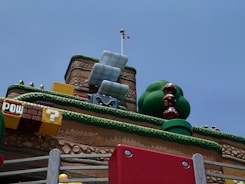 A whimsical, colorful landscape reminiscent of a video game environment. Large, stacked brown mushroom-like characters with big eyes stand next to a sculpture of a green bush. Various blocks and elements are inspired by classic platformer aesthetics, including a yellow block with a question mark and a brown block with 'POW' written on it. A red and white flag is placed atop a platform resembling a fortress with tiered layers covered in faux grass.