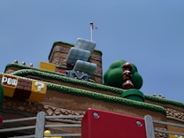 A whimsical, colorful landscape reminiscent of a video game environment. Large, stacked brown mushroom-like characters with big eyes stand next to a sculpture of a green bush. Various blocks and elements are inspired by classic platformer aesthetics, including a yellow block with a question mark and a brown block with 'POW' written on it. A red and white flag is placed atop a platform resembling a fortress with tiered layers covered in faux grass.