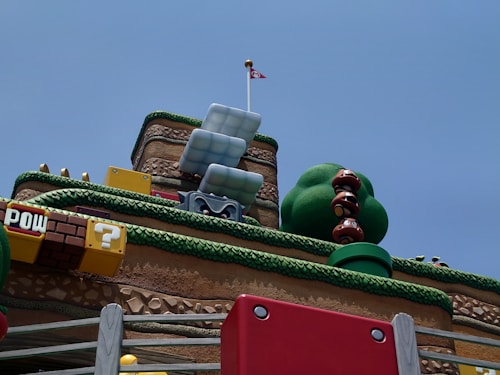 A whimsical, colorful landscape reminiscent of a video game environment. Large, stacked brown mushroom-like characters with big eyes stand next to a sculpture of a green bush. Various blocks and elements are inspired by classic platformer aesthetics, including a yellow block with a question mark and a brown block with 'POW' written on it. A red and white flag is placed atop a platform resembling a fortress with tiered layers covered in faux grass.