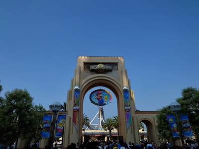An entrance archway with a prominent sign for Universal Studios. Banners with colorful designs featuring animated characters are displayed on both sides. A large circular emblem with an animated character is centered above the arch. In the background, there are palm trees and part of a roller coaster structure.