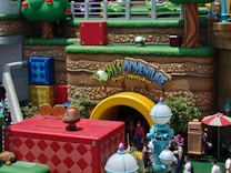 A vibrant, colorful theme park area with large, cartoonish structures resembling a video game landscape. The scene includes oversized blocks in blue, red, and yellow, with a large arch labeled 'Adventure' and characters dressed up in costumes. There are whimsical trees and decor resembling mushrooms, with guests walking around soaking up the fun atmosphere.