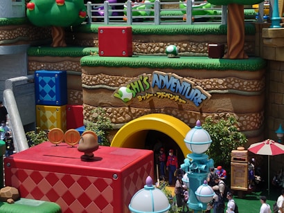 A vibrant, colorful theme park area with large, cartoonish structures resembling a video game landscape. The scene includes oversized blocks in blue, red, and yellow, with a large arch labeled 'Adventure' and characters dressed up in costumes. There are whimsical trees and decor resembling mushrooms, with guests walking around soaking up the fun atmosphere.