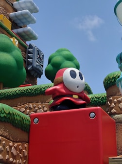 A large cartoonish character dressed in red is standing on a red block. The background features whimsical, artificial greenery, a stone wall pattern, and stacked, cloud-like structures. A menacing, grey stone face with red eyes is visible on the left side.