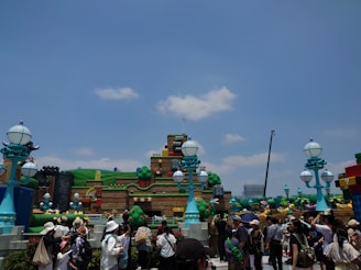 A vibrant outdoor amusement park setting featuring colorful, whimsical, and large structures resembling a video game-themed environment. There are numerous people walking around and observing the attractions. The sky is clear with scattered clouds.