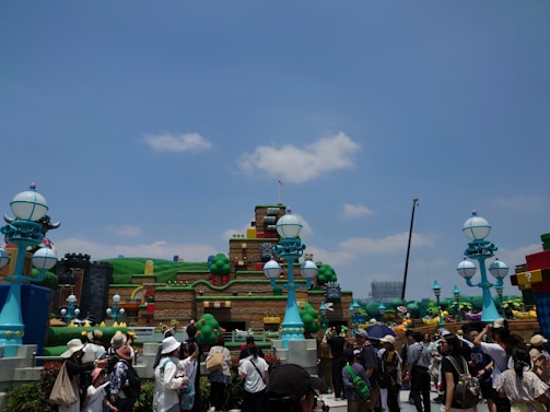 A vibrant outdoor amusement park setting featuring colorful, whimsical, and large structures resembling a video game-themed environment. There are numerous people walking around and observing the attractions. The sky is clear with scattered clouds.
