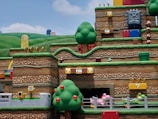 A vibrant and colorful theme park scene depicting a section of an amusement attraction themed around a popular video game. The landscape features stylized hills, trees, and stacked platforms typical of the game’s aesthetic. There are recognizable game elements such as question mark blocks, brick platforms, gold coins, and whimsical character figures like Goombas and a Thwomp block. Various interactive elements and roller coaster tracks are also visible, creating an immersive and playful atmosphere.