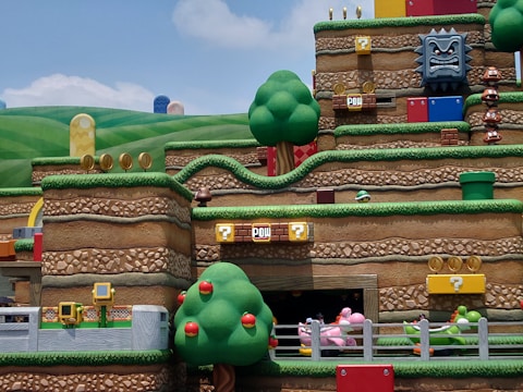 A vibrant and colorful theme park scene depicting a section of an amusement attraction themed around a popular video game. The landscape features stylized hills, trees, and stacked platforms typical of the game’s aesthetic. There are recognizable game elements such as question mark blocks, brick platforms, gold coins, and whimsical character figures like Goombas and a Thwomp block. Various interactive elements and roller coaster tracks are also visible, creating an immersive and playful atmosphere.
