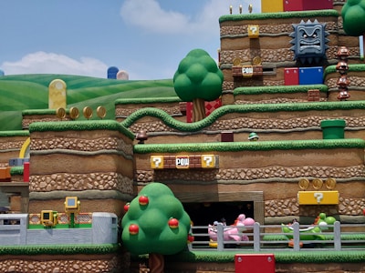 A vibrant and colorful theme park scene depicting a section of an amusement attraction themed around a popular video game. The landscape features stylized hills, trees, and stacked platforms typical of the game’s aesthetic. There are recognizable game elements such as question mark blocks, brick platforms, gold coins, and whimsical character figures like Goombas and a Thwomp block. Various interactive elements and roller coaster tracks are also visible, creating an immersive and playful atmosphere.