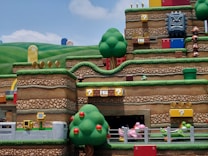 A vibrant and colorful theme park scene depicting a section of an amusement attraction themed around a popular video game. The landscape features stylized hills, trees, and stacked platforms typical of the game&rsquo;s aesthetic. There are recognizable game elements such as question mark blocks, brick platforms, gold coins, and whimsical character figures like Goombas and a Thwomp block. Various interactive elements and roller coaster tracks are also visible, creating an immersive and playful atmosphere.