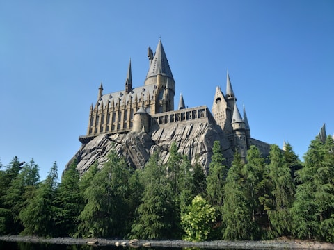 A grand castle sits majestically atop a rocky hill, surrounded by lush green trees under a clear blue sky. The castle displays a series of tall spires, turrets, and intricate architectural details, indicative of a medieval style.
