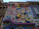 A colorful mural features animated characters resembling small yellow creatures with goggles, overalls, and playful expressions. They are depicted climbing on a ladder against a vibrant backdrop with abstract graffiti patterns and whimsical elements. The scene includes decorative architectural elements surrounding the artwork, adding a playful and imaginative atmosphere.