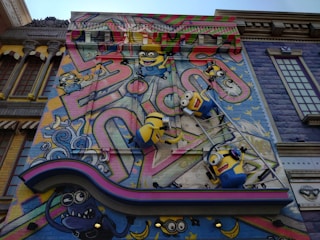 A colorful mural features animated characters resembling small yellow creatures with goggles, overalls, and playful expressions. They are depicted climbing on a ladder against a vibrant backdrop with abstract graffiti patterns and whimsical elements. The scene includes decorative architectural elements surrounding the artwork, adding a playful and imaginative atmosphere.
