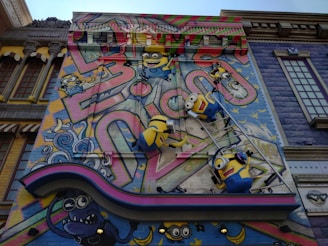 A colorful mural features animated characters resembling small yellow creatures with goggles, overalls, and playful expressions. They are depicted climbing on a ladder against a vibrant backdrop with abstract graffiti patterns and whimsical elements. The scene includes decorative architectural elements surrounding the artwork, adding a playful and imaginative atmosphere.