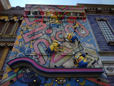A colorful mural features animated characters resembling small yellow creatures with goggles, overalls, and playful expressions. They are depicted climbing on a ladder against a vibrant backdrop with abstract graffiti patterns and whimsical elements. The scene includes decorative architectural elements surrounding the artwork, adding a playful and imaginative atmosphere.