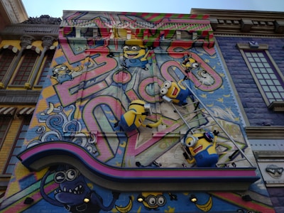A colorful mural features animated characters resembling small yellow creatures with goggles, overalls, and playful expressions. They are depicted climbing on a ladder against a vibrant backdrop with abstract graffiti patterns and whimsical elements. The scene includes decorative architectural elements surrounding the artwork, adding a playful and imaginative atmosphere.
