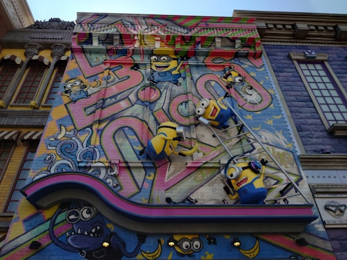 A colorful mural features animated characters resembling small yellow creatures with goggles, overalls, and playful expressions. They are depicted climbing on a ladder against a vibrant backdrop with abstract graffiti patterns and whimsical elements. The scene includes decorative architectural elements surrounding the artwork, adding a playful and imaginative atmosphere.