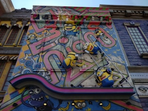 A colorful mural features animated characters resembling small yellow creatures with goggles, overalls, and playful expressions. They are depicted climbing on a ladder against a vibrant backdrop with abstract graffiti patterns and whimsical elements. The scene includes decorative architectural elements surrounding the artwork, adding a playful and imaginative atmosphere.