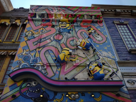 A colorful mural features animated characters resembling small yellow creatures with goggles, overalls, and playful expressions. They are depicted climbing on a ladder against a vibrant backdrop with abstract graffiti patterns and whimsical elements. The scene includes decorative architectural elements surrounding the artwork, adding a playful and imaginative atmosphere.