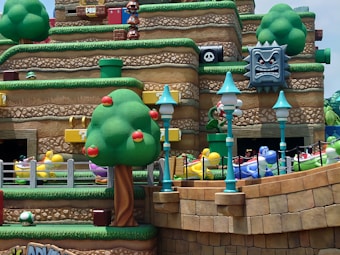 The scene features a vibrant and colorful amusement park setting inspired by a popular video game. It includes stylized green hills with layered textures, large cartoon trees adorned with red fruits, and floating blocks with question marks. Characters resembling dinosaurs in various colors, including yellow, blue, green, and purple, appear on a ride surrounded by pathways and fences. The area is decorated with whimsical street lamps and a large, menacing stone face with spikes.