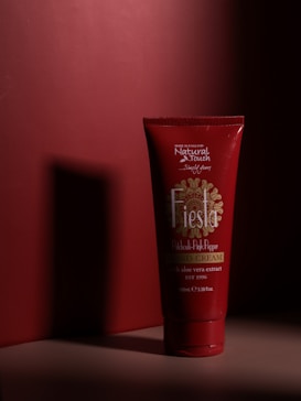 A red tube of hand cream labeled 'Fiesta' is standing upright against a matching red background. The packaging design features intricate patterns and text in white and gold, highlighted by a dramatic shadow and light effect, creating a stark contrast.