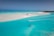 A serene tropical beach with clear turquoise waters and white sandy shores. A person is kiteboarding, leaving a trail in the water, and the kite is brightly colored against the sky. The setting is peaceful and expansive, with a horizon that stretches across the tranquil sea.