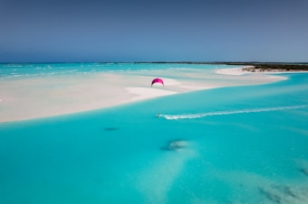 A serene tropical beach with clear turquoise waters and white sandy shores. A person is kiteboarding, leaving a trail in the water, and the kite is brightly colored against the sky. The setting is peaceful and expansive, with a horizon that stretches across the tranquil sea.