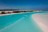 A scenic view of a meandering turquoise water channel surrounded by white sandy beaches and sparse vegetation. A kite surfer is gliding across the water, creating a trail behind. The sky is clear with a rich blue tone, adding to the tranquil atmosphere.
