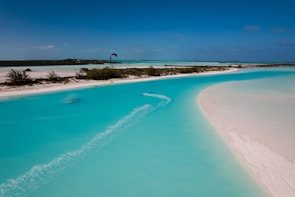 A scenic view of a meandering turquoise water channel surrounded by white sandy beaches and sparse vegetation. A kite surfer is gliding across the water, creating a trail behind. The sky is clear with a rich blue tone, adding to the tranquil atmosphere.