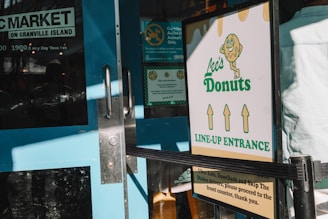 A doorway with a teal blue door featuring glass panes and metal handles. Nearby, a sign for Lee's Donuts indicates the line-up entrance, with graphic design elements including a donut character and directional arrows. Sunlight casts shadows across the scene, reflecting a bustling market environment.
