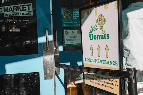 A doorway with a teal blue door featuring glass panes and metal handles. Nearby, a sign for Lee's Donuts indicates the line-up entrance, with graphic design elements including a donut character and directional arrows. Sunlight casts shadows across the scene, reflecting a bustling market environment.