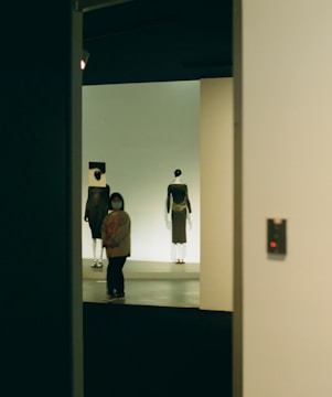 A dimly lit room features a person wearing a mask walking past two mannequins dressed in modern clothing. The mannequins are displayed against a simple backdrop, and the scene is framed by a doorway.