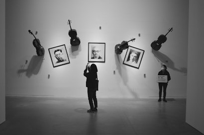 A gallery setting where several violins are mounted on the wall alongside framed black and white portraits. A person is taking a photo with a phone while another person stands holding a sign.