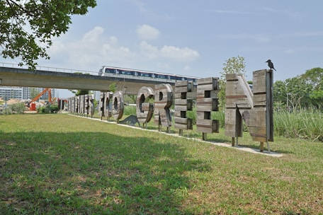 A wooden sign with the words 'Tampines Eco Green' is placed on a grassy field. A bird is perched on the last letter of the sign. In the background, there is an elevated train track with a train passing by. Buildings, construction equipment, and trees are also visible.