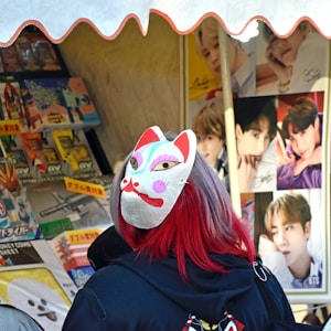 A person with vibrant red and purple hair is wearing a traditional Japanese fox mask. They are facing shelves filled with various merchandise, including posters with celebrities and toys. The setting seems to be an open market or a stall under a canopy.