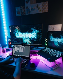 A dimly lit room features a gaming setup with two monitors displaying the word 'LEGION' in bright neon. A person is working on a laptop with the keyboard illuminated by soft blue and purple ambient lighting. The desk is cluttered with accessories, and in the background, there are posters and LED strip lights enhancing the atmosphere.