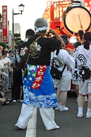 A person wearing a colorful traditional garment with a blue and red design is standing among a group of people. They carry a decorated horn. In the background, there is a large drum being played, surrounded by others in similar traditional attire.