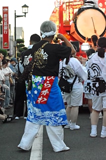 A person wearing a colorful traditional garment with a blue and red design is standing among a group of people. They carry a decorated horn. In the background, there is a large drum being played, surrounded by others in similar traditional attire.