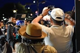 A crowd of people is gathered at an outdoor event during the night. Several individuals are capturing photos and videos using smartphones and tablets. The crowd is diverse, indicating a busy and popular event. The night setting is highlighted by the dark sky and artificial lights illuminating the scene.
