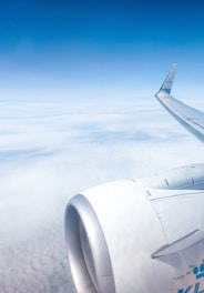 An airplane wing and engine are visible against a backdrop of a clear blue sky and a layer of clouds below. The tip of the wing features branding from an airline, suggesting that the aircraft is mid-flight at a high altitude.