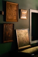 Several framed art pieces are displayed on a dark green wall. The frames are ornate with classic designs, and one of the paintings appears to have historical or classical themes. A large mirror with a simple white frame is also visible.