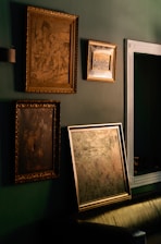 Several framed art pieces are displayed on a dark green wall. The frames are ornate with classic designs, and one of the paintings appears to have historical or classical themes. A large mirror with a simple white frame is also visible.
