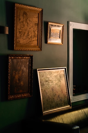Several framed art pieces are displayed on a dark green wall. The frames are ornate with classic designs, and one of the paintings appears to have historical or classical themes. A large mirror with a simple white frame is also visible.