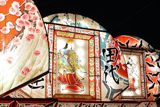 Brightly illuminated traditional Japanese artworks are displayed, featuring intricate designs and vibrant colors. The central piece showcases a figure in traditional attire, surrounded by ornate patterns and calligraphy. Additional panels include floral motifs and dynamic scenes with intricate details.