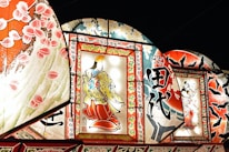 Brightly illuminated traditional Japanese artworks are displayed, featuring intricate designs and vibrant colors. The central piece showcases a figure in traditional attire, surrounded by ornate patterns and calligraphy. Additional panels include floral motifs and dynamic scenes with intricate details.