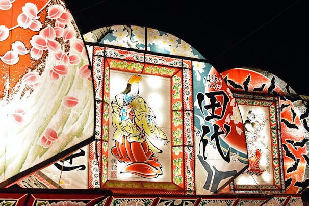 Brightly illuminated traditional Japanese artworks are displayed, featuring intricate designs and vibrant colors. The central piece showcases a figure in traditional attire, surrounded by ornate patterns and calligraphy. Additional panels include floral motifs and dynamic scenes with intricate details.
