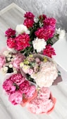 A collection of vibrant floral arrangements featuring pink peonies, white peonies, and a variety of other blossoms in different colors. The flowers are neatly arranged in bouquets placed on a light wooden floor with a textured grey wall in the background.