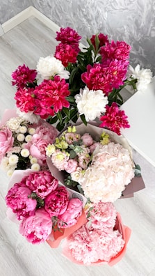 A collection of vibrant floral arrangements featuring pink peonies, white peonies, and a variety of other blossoms in different colors. The flowers are neatly arranged in bouquets placed on a light wooden floor with a textured grey wall in the background.