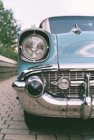 The image features a close-up view of the front of a vintage car, focusing on the headlight and chrome details. The car's blue body contrasts with the shiny, reflective chrome bumper. The setting appears to be outdoors, on a cobblestone path with green foliage in the background.