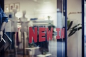 Red letters spelling 'NEW IN' are prominently displayed on a glass window, hinting at the entrance to a retail store. Inside, mannequins and display racks featuring clothing items can be seen, indicating a fashion shop setup. Blurred elements and reflections add depth to the scene.