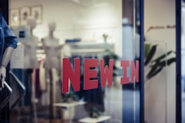 Red letters spelling 'NEW IN' are prominently displayed on a glass window, hinting at the entrance to a retail store. Inside, mannequins and display racks featuring clothing items can be seen, indicating a fashion shop setup. Blurred elements and reflections add depth to the scene.